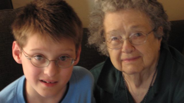 A young boy with glasses in a blue shirt sits next to an older woman with curly gray hair and glasses wearing a dark green top.