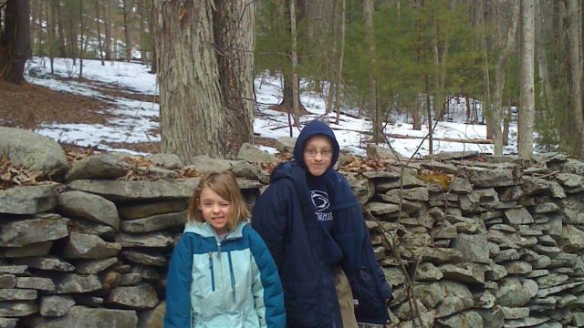 Two children pose in front of a dry stone wall in a forest setting during winter. Two children in winter jackets standing in front of a stone wall in a wooded area with snow on the ground.