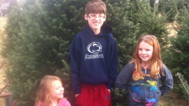 Three children standing in front of a large Christmas tree at a tree farm.