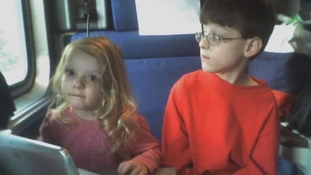 Two children seated together on a train. Two young children sitting on a train, one girl with blonde hair and pink shirt looking at camera, one boy in red shirt and glasses looking away.