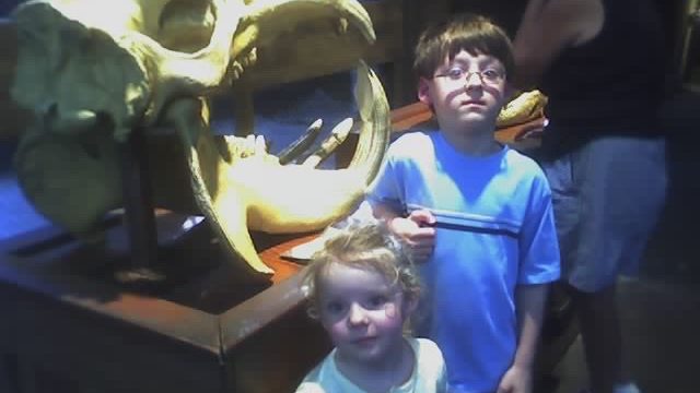 Two children pose for a photo in front of a large animal skull exhibit. Two young children standing in front of a large animal skull display at what appears to be a museum or exhibit.