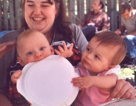 A smiling woman holds two babies; one baby holds a large white circular object in front of his face.