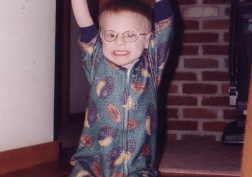 A young child wearing glasses and a patterned onesie stands on wooden steps with arms raised in the air.