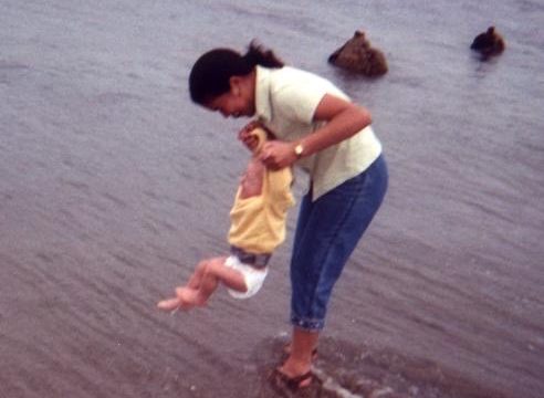 Woman holding a child near the shoreline. A woman in a yellow shirt and blue jeans holds a small child in white shorts at the water's edge on a beach.