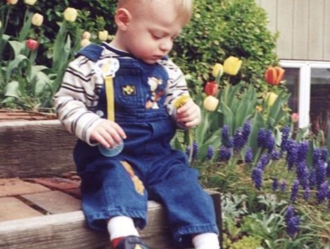 A young child with blond hair wearing striped shirt and blue overalls sitting on wooden steps in a garden holding small yellow object.