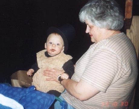 An older woman with gray hair holds a baby wearing a top hat and beige outfit.