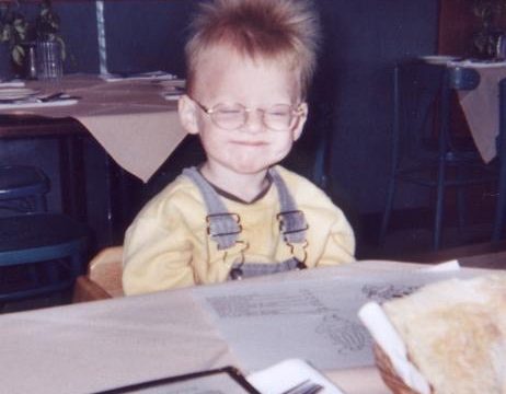 A young child smiles while seated at a restaurant table. A young child with spiky hair and glasses sits at a restaurant table wearing a yellow shirt with suspenders.