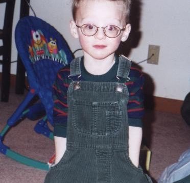 A young boy wearing round glasses and green overalls sits in front of a blue baby seat.