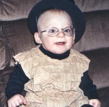 A smiling baby wearing a black hat, round glasses, and a beige textured vest over a black long-sleeve shirt sitting on a brown couch.