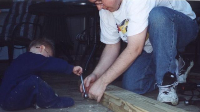 A man in a white t-shirt and jeans kneels on a wooden floor, holding a metal tool while a small child in blue clothing sits nearby.