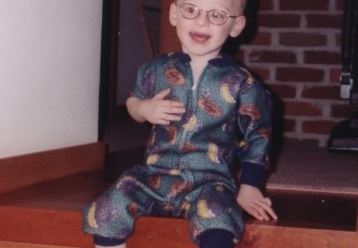 A young child wearing glasses and a patterned onesie sits on wooden steps indoors.