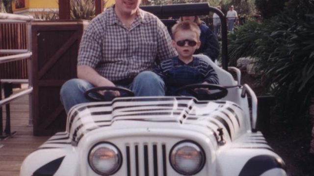 A father and son enjoy a ride together in a themed vehicle. A man and a young boy wearing sunglasses sit in a white striped ride vehicle at an amusement park.