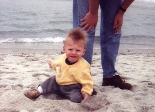 Child sitting on the sand with an adult standing behind. A young child in a yellow sweater sits on the beach while an adult stands nearby.