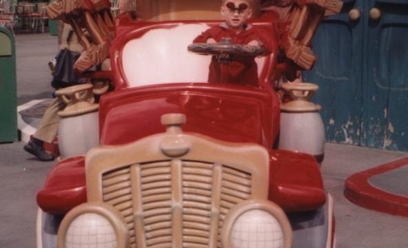 Child seated in a colorful, themed ride vehicle at an amusement park. A child sits in a red vintage-style ride vehicle with oversized props and decorative elements.