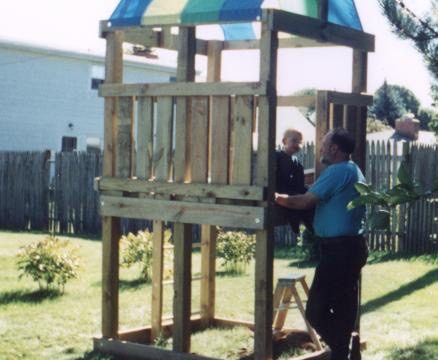 Two men standing beside a wooden playhouse with a striped roof in a backyard.