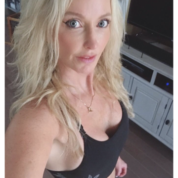 A woman with blonde hair wearing a black sports bra taking a selfie in a room with a TV and wooden furniture.