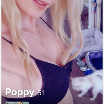 A woman with blonde hair wearing a black top, smiling at the camera, with a small white cat walking nearby in a room.