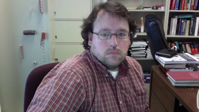 A man in a plaid shirt sits in an office chair, looking directly at the camera. A man with glasses and a plaid shirt sits in an office chair, looking directly at the camera.