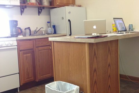 A home office setup in a kitchen featuring two laptops on a counter. A kitchen with wooden cabinets, a white refrigerator, and two laptops on a counter.