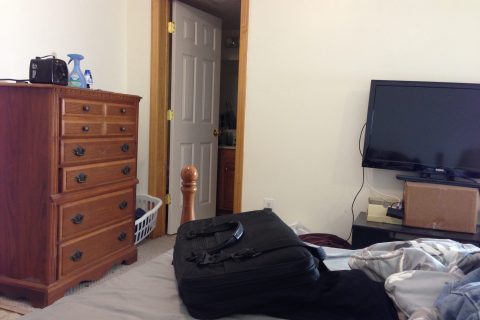 A tidy bedroom featuring a wooden dresser, flat-screen TV, and a black suitcase on the bed. A bedroom with a dresser, TV, suitcase on bed, and open door to another room.